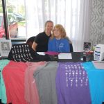 Jeri Cooper sitting at exhibitor table where tshirts of different colors are draped across the table.